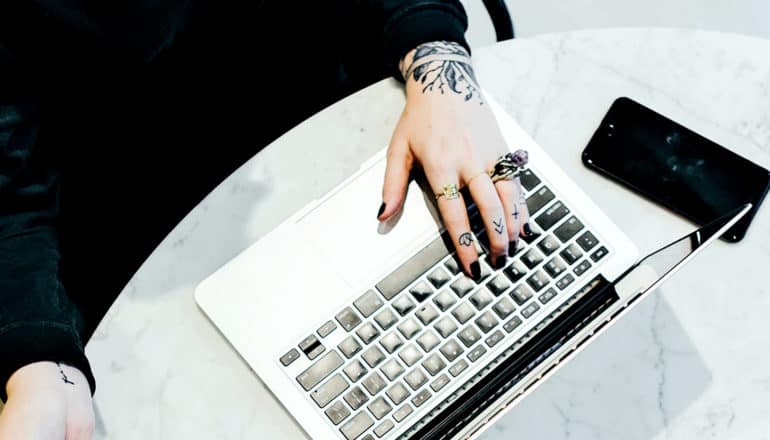 A person with hand tattoos uses a laptop that's sitting on a marble table with a phone nearby