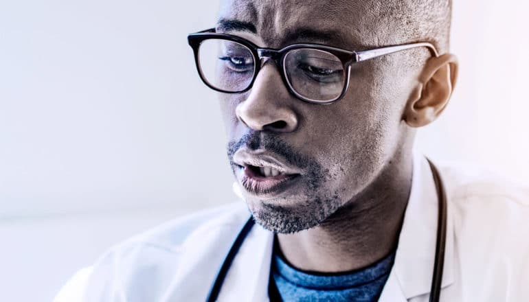 A doctor wearing glasses looks down with focus or concern as he talks to his patient