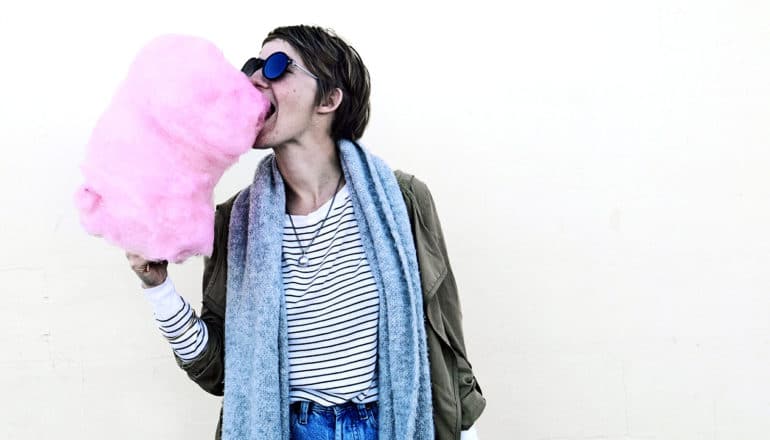 A person in sunglasses and a long scarf eats a large mass of pink cotton candy