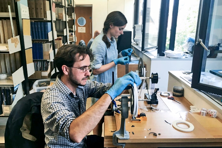 Two researchers with blue gloves on feed film into reels on a table, one standing, one sitting.