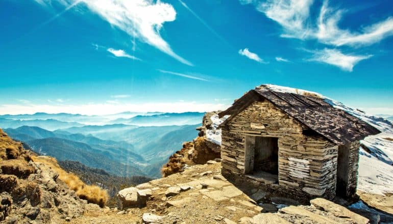 stone hut sits at edge of cliff overlooking Himalayan mountains and blue sky
