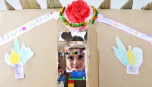 child peeks through cardboard structure for Sukkot with decorations and "happy sukkot" signs