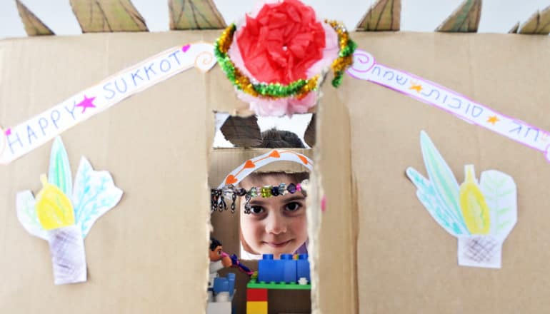 child peeks through cardboard structure for Sukkot with decorations and "happy sukkot" signs