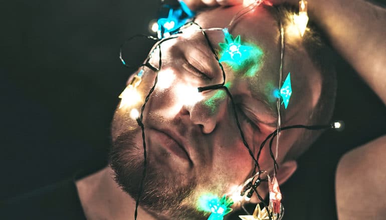 A man holds string lights over his face, dangling down from the top of his head, on a black background
