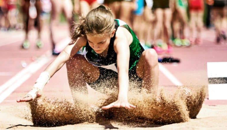 A long jumper lands in the sand while steadying herself, with a crowd of other student athletes behind her on the track