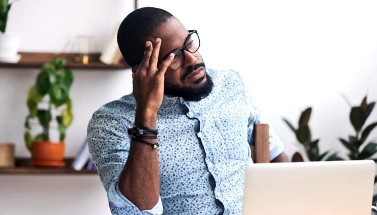 A man with glasses sits at a desk, looking off while resting his head on his hand