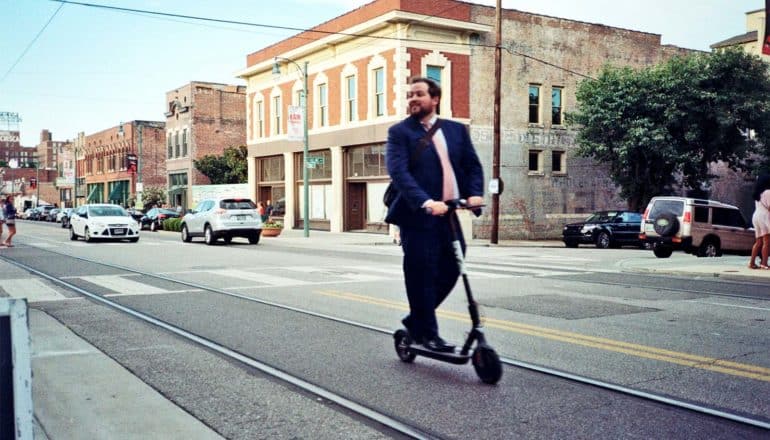A man in a suit rides an electric scooter down the street, turning to smile at pedestrians, as cars pass buildings in the background