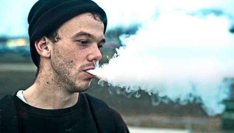 A man vaping breathes out a large cloud while wearing a black sweater and beanie