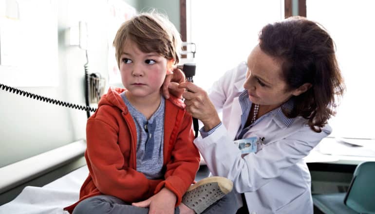 serious, flushed child sits on examining table, doctor looks into ear with otoscope