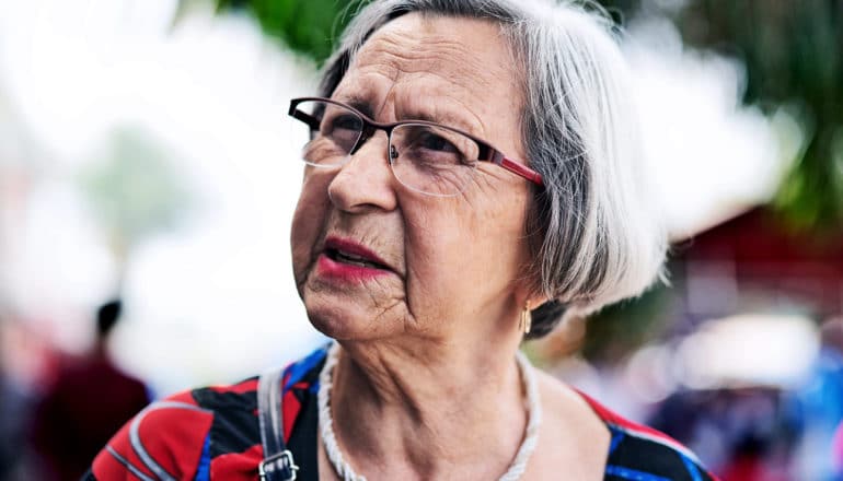 An older woman with white hair and glasses walks with a confused look on her face