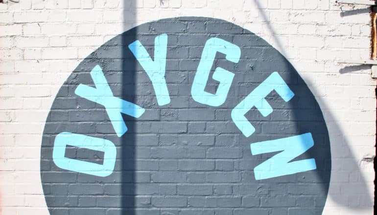 A mural on a white brick wall has a dark circle with the word "oxygen" in blue letters inside