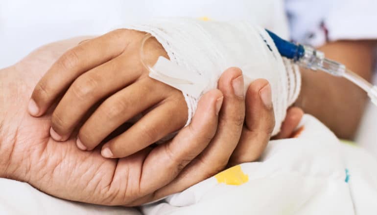 A parent holds a child's bandaged hand, which has an IV going into it