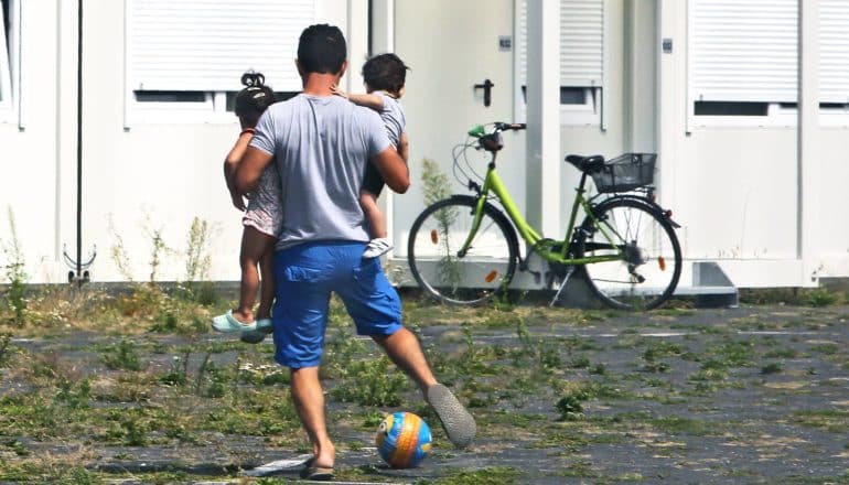 man carries two toddlers while kicking ball in courtyard