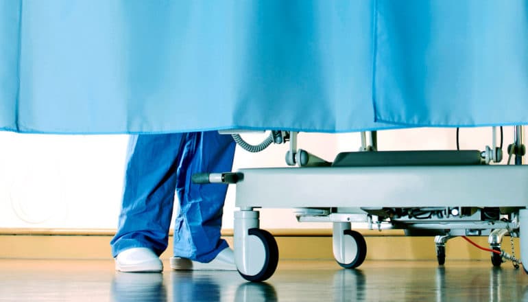 A patient's bed sits behind a blue curtain in a hospital with a scrubs-clad specialist at the bedside