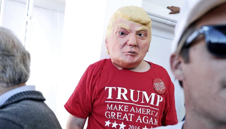 A person in a Make America Great Again t-shirt wears a rubber Trump mask