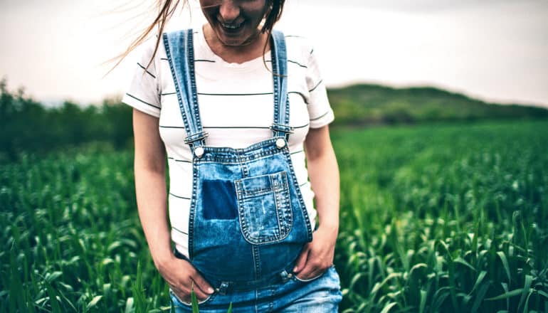 a pregnant woman walks with her hands in her overall pockets through a field of tall grass