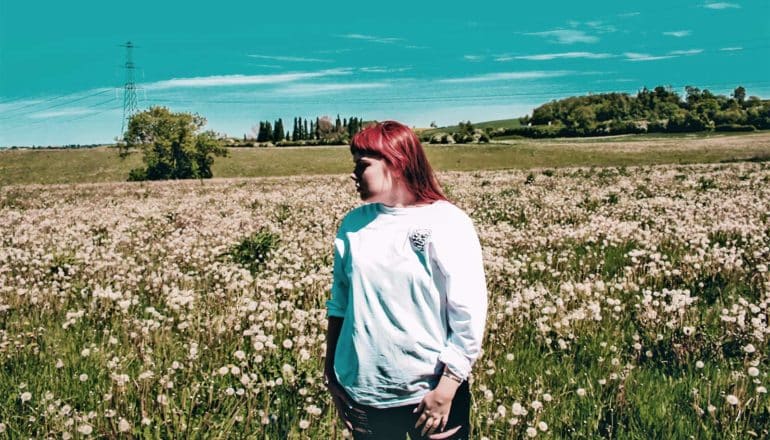 A young red-haired woman stands in large open field covered in flowers while wearing a white shirt, with the bright blue sky in the background