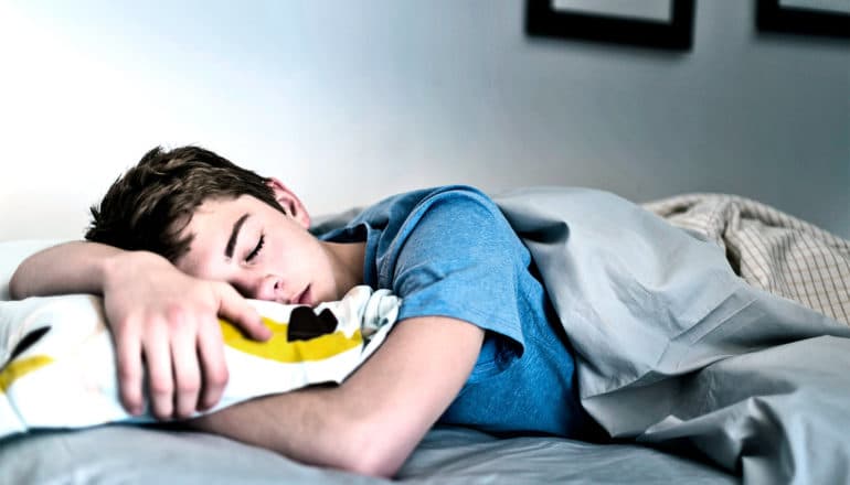 A teen boy sleeps on his stomach with his arms over and under his pillow