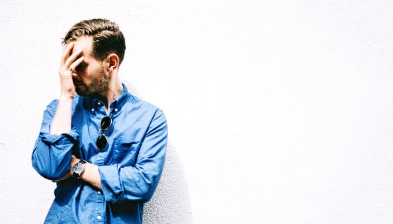 A man leans against a white wall with his hand over his face and his other arm crossed