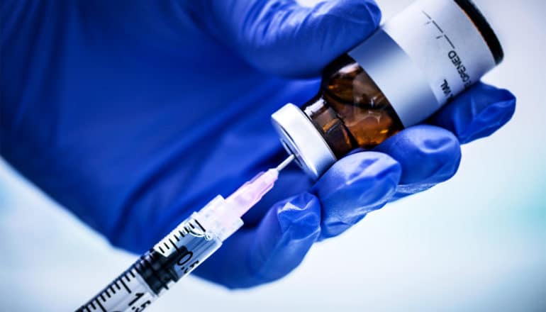 A blue-gloved hand holds a vaccine bottle as a syringe extracts the vaccine for injection