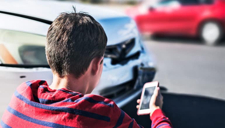 A teen in a red shirt looks at their phone while sitting on the curb, ready to call, at the scene of a car accident
