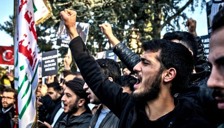 Men raise their fists while dressed mostly in black to protest Saudi Arabia, while other protestors hold signs and flags