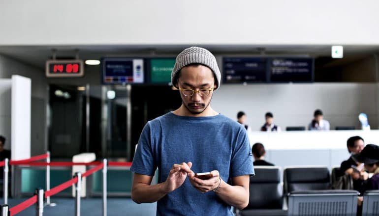 A man in a beanie and t-shirt looks down at his phone while he's standing in an airport gate