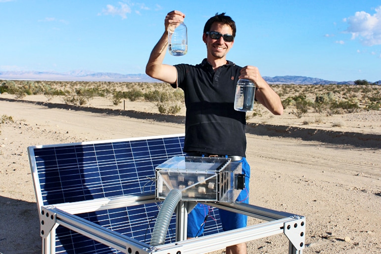 Prévot stands next to the water harvester and solar panel in the desert while holding up two bottles of water. He's grinning widely while wearing a black shirt and sunglasses.