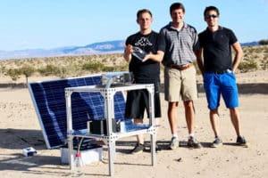Three researchers stand in the desert near the water harvester setup, one of them holding a bottle of water