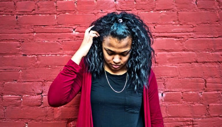 A young woman moves her hair out of her face while standing against a red brick wall