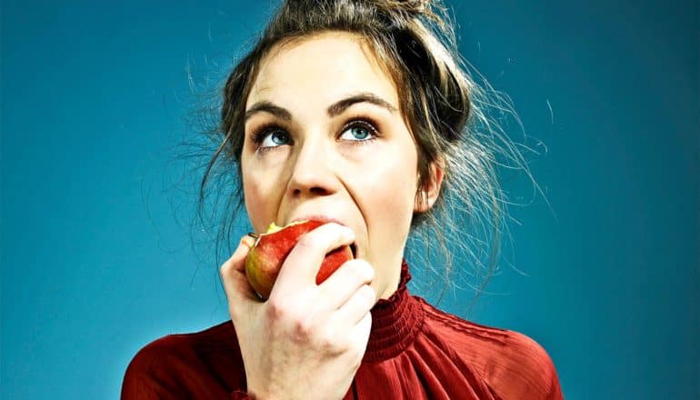 A woman in red eats a red apple while looking upwards against a blue background
