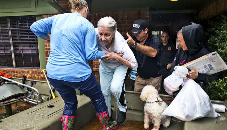 An older woman gets help to leave her house during a flood, as people help her into a boat and her dog watches