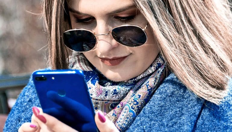 A woman with a blue coat and sunglasses on texts on a blue phone while sitting on a bench