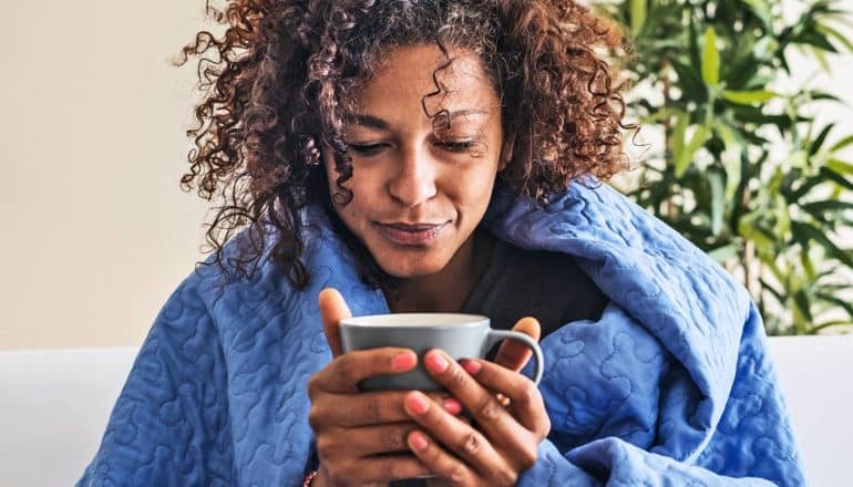 A woman sits with a blue blanket wrapped around her shoulders as she looks down at a cup of tea