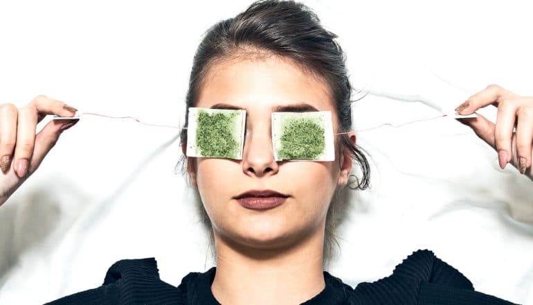 A young woman has green tea bags over her eyes as she lies in bed