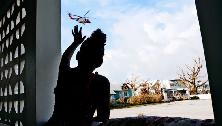 A young girl reaches up out of a window towards a rescue helicopter while in the background there are houses that have been damaged in the hurricane and a flooded street
