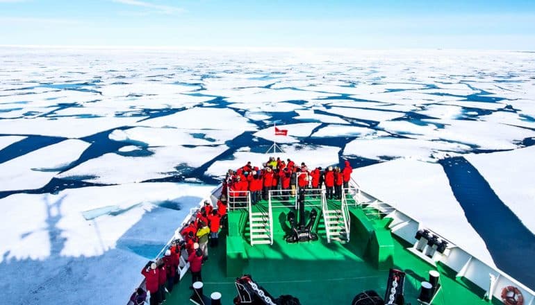 a green ship with passengers in red jackets moves through Arctic sea ice, which is broken into large sheets with the ocean showing in between