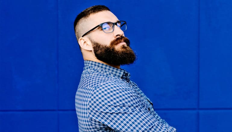 A man with a beard and glasses crosses his arms and looks over his shoulder while standing against a blue wall
