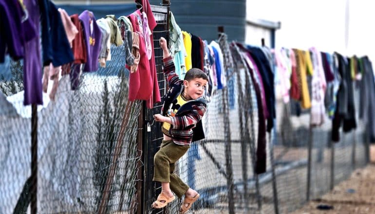 A young Syrian child hangs off a fence covered in clothes in a refugee camp