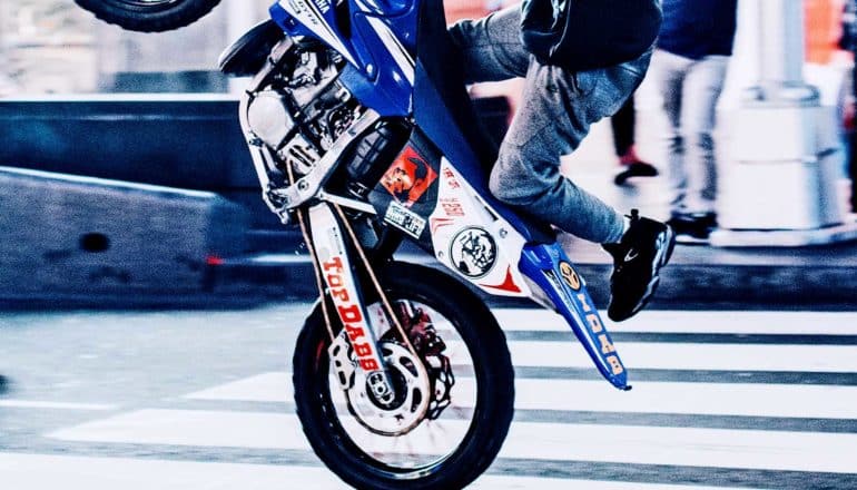 A person on a dirt bike does a wheelie on the street as they pass over a crosswalk