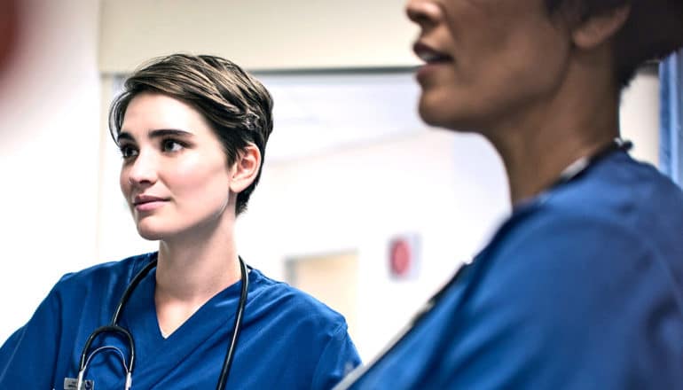 Two doctors in blue scrubs stand in a circle in a hospital listening