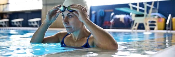 A woman with goggles and a swim cap stands in a swimming pool with light coming in windows from outside