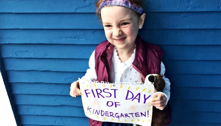 A kindergartener holds up a sign that says "First day of kindergarten" as she smiles and stands in front of a blue wall while posing for a picture