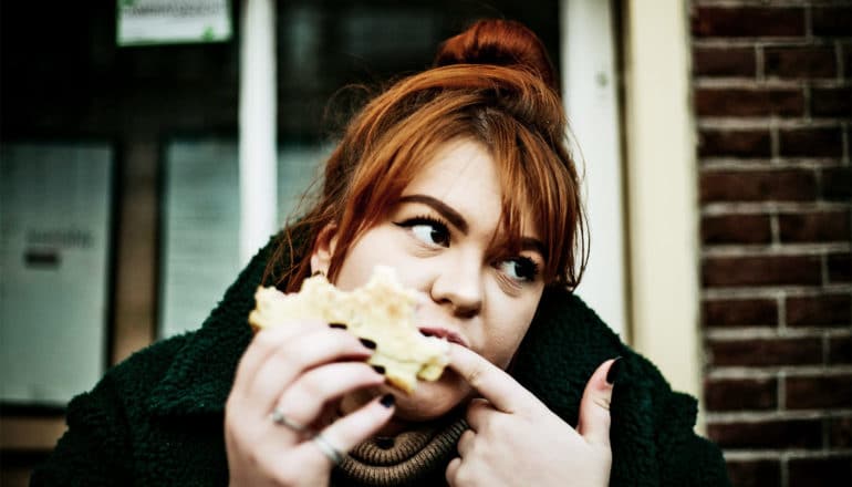 A woman eating a sandwich on white bread licks her finger while looking to her left