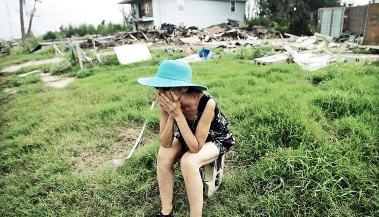 A woman sits on a cinderblock in front of a destroyed home with her head in her hands