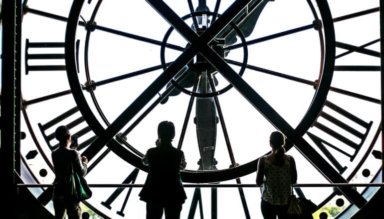 3 people stand behind the face of a large clock, looking out through its face