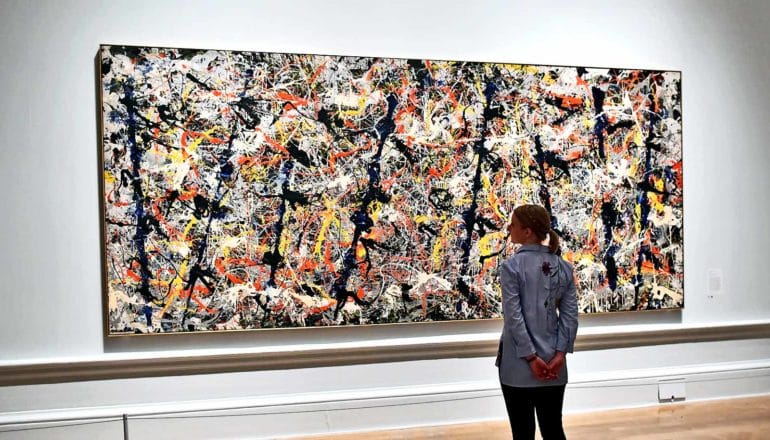 A woman looks up at a Jackson Pollock painting in a museum, which has black, orange, yellow, and white paint splashes