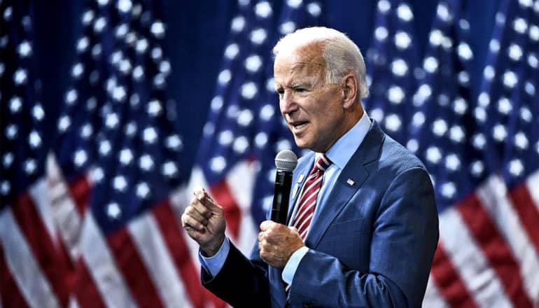 Joe Biden speaks into a microphone while holding one hand up and standing in front of a row of American flags