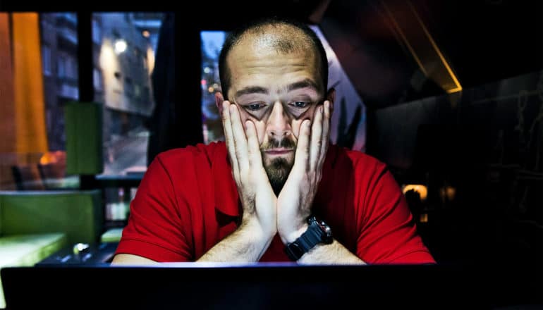 A man has both hands on his cheeks as he looks down at a screen with a blank face