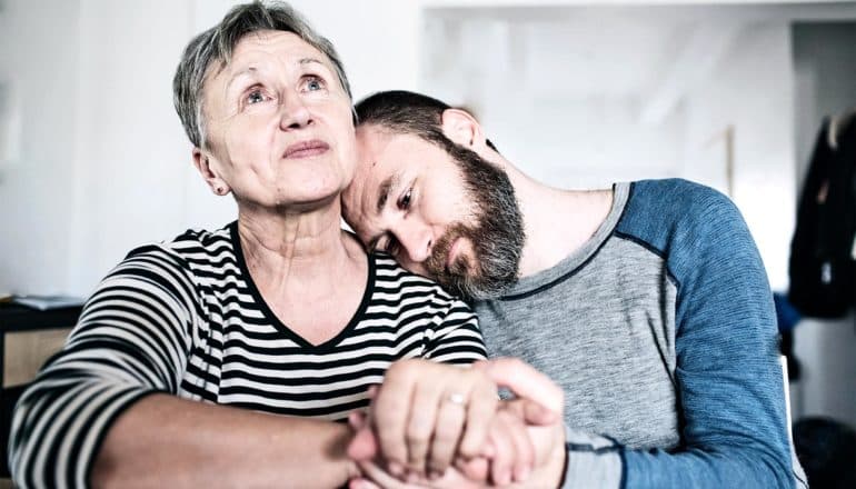 A mother and her son sit at a table holding hands with his head on her shoulder